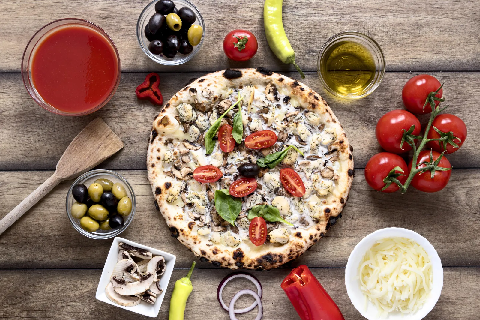 A wood-fired pizza surrounded by ingredients: cherry tomatoes, olives, cheese, mushrooms, tomato sauce, olive oil, and peppers on a wooden table.